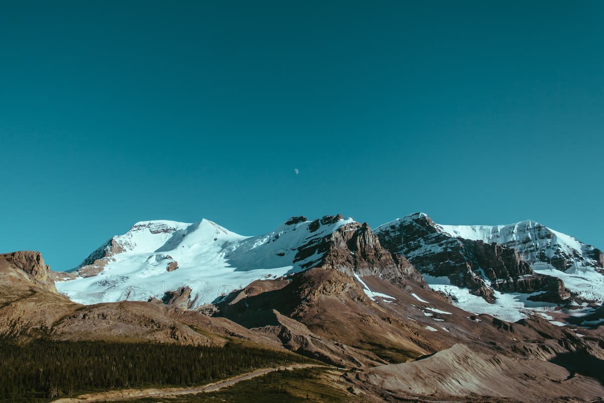 A snow-covered mountain range under a clear sky, with the moon still up.